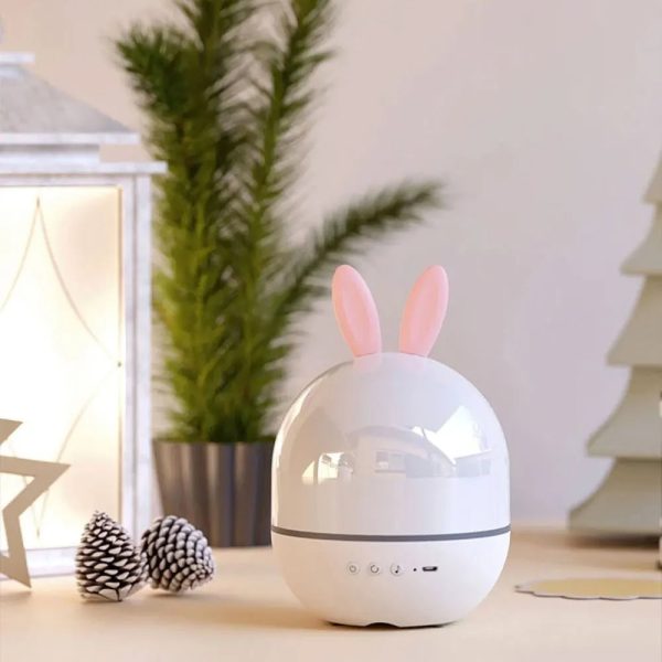 Projetor de galáxia com forma de ovo e orelhas de coelho, em ambiente decorativo com plantas e objetos de decoração.