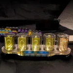 Relógio de números digitais com tubos de plasma iluminados em cores diferentes, exibindo a hora 20:24:21, colocado sobre uma superfície escura com teclado de computador ao fundo.