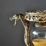 Detalhe de uma cobra dourada de metal com cabeça levantada, envolta num recipiente de vidro com tampa decorativa dourada, fundo cinzento.