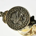 Detalhe de uma peça de joalharia de metal com símbolo de dragão, decorada com padrão de nós celtas ao redor, vista de perto.