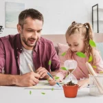Pai e filha a realizar um experimento científico com um vaso, areia, uma ampulheta e folhas verdes, numa sala bem iluminada.