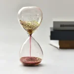 Relógio de areia transparente com glitter dourado, mostrando o fluxo de areia de cima para baixo, com livros ao fundo.