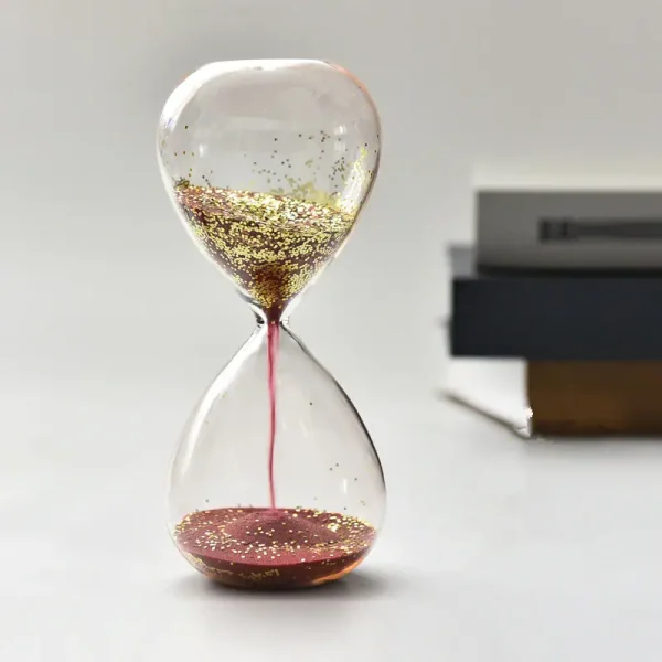 Relógio de areia transparente com glitter dourado, mostrando o fluxo de areia de cima para baixo, com livros ao fundo.