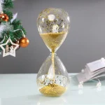 Ampulheta decorativa com areia dourada e glitter, sobre mesa branca, com decoração de Natal ao fundo, incluindo árvore com enfeites.