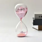 Relógio de areia transparente com areia rosa e partículas brilhantes, em fundo branco, com livros ao lado.