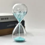 Relógio de areia transparente com areia azul em movimento, fundo branco, livros ao lado, usado para demonstrações científicas.