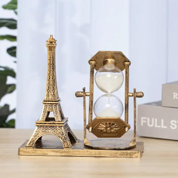 Relógio de areia dourado com estrutura decorativa em forma de Torre Eiffel ao lado, colocado numa base de madeira, com fundo de cortina branca e caixas de armazenamento.