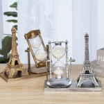 Relógio de areia de vidro com estrutura metálica, acompanhado por duas miniaturas da Torre Eiffel em diferentes cores, decorando uma mesa de madeira.