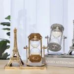 Relógio de areia dourado com base de vidro, ao lado de uma réplica da Torre Eiffel em metal dourado e uma outra em prata, em fundo de painéis brancos e uma planta verde.