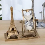 Relógio de areia com estrutura metálica dourada ao lado de uma miniatura da Torre Eiffel de metal dourado, ambos em uma base de madeira.