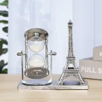 Relógio de areia prateado ao lado de uma miniatura da Torre Eiffel em uma base de madeira, com fundo de cortina branca e livros ao lado.