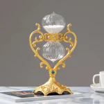 Relógio de areia com estrutura dourada ornamentada e detalhes azuis, colocado sobre uma mesa com revistas e uma chávena branca ao lado.