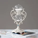 Relógio de areia decorativo com estrutura de metal ornamentada e vidro transparente, colocado sobre uma revista com óculos de leitura ao lado.