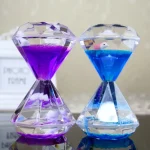 Relógio de areia com cristais transparentes, um com areia roxa e outro com areia azul, ambos com detalhes brilhantes e pequenos elementos decorativos internos.