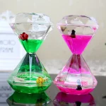 Dois relógios de areia de vidro, um com areia verde e peixes vermelhos, outro com areia rosa e peixes pretos, ambos em cima de uma superfície refletora.