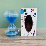 Relógio de areia de vidro com líquido azul e base transparente, ao lado de uma caixa decorativa com buracos e uma janela oval escura.