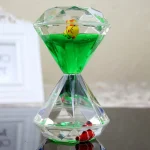 Relógio de areia com estrutura de cristal, preenchido com líquido verde e pequenos brinquedos de plástico coloridos dentro.