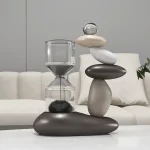 Escultura moderna composta por pedras de diferentes tamanhos e cores, com um elemento de vidro transparente no topo, colocada numa mesa branca.