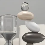 Conjunto de pedras lisas de cores branca, cinza, preta empilhadas com esfera de vidro transparente no topo, ao lado de um recipiente de vidro com líquido.