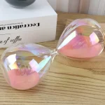 Ampulheta de vidro transparente com areia rosa, colocada numa superfície de madeira, com caixa branca ao fundo.