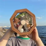 Caixa de madeira hexagonal com folhas, pedra e elementos decorativos, vista de frente com fundo de mar e céu azul.