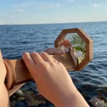 Espelho de mão com moldura de madeira octogonal, refletindo folhas verdes e uma flor, com vista para o mar ao fundo.