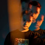 Fotografia de um rapaz com efeito de duplicação de rosto, mostrando várias camadas e ângulos, com iluminação dramática e fundo escuro.