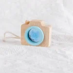 Câmara de brinquedo de madeira com lente azul e detalhes em madeira natural, colocada sobre tecido branco, com cordão de puxar ao lado.