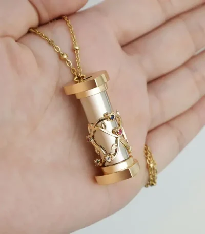 Colar dourado com pingente de cilindro prateado decorado com uma figura de árvore e pedras coloridas, segurado na mão.