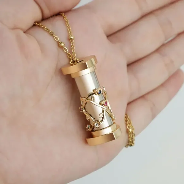 Colar dourado com pingente de cilindro prateado decorado com uma figura de árvore e pedras coloridas, segurado na mão.