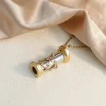 Colar com pingente de microscópio dourado, detalhe de lente e base, sobre fundo de tecido de seda bege.
