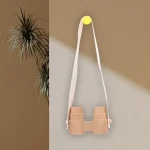 Binóculos de madeira decorativos suspensos por uma correia de tecido branco, fixados numa parede de cores diferentes, com uma planta ao lado.