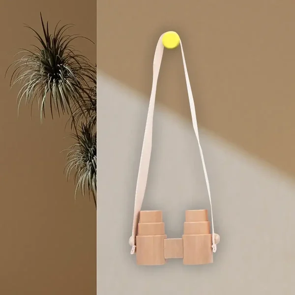 Binóculos de madeira decorativos suspensos por uma correia de tecido branco, fixados numa parede de cores diferentes, com uma planta ao lado.