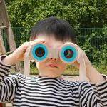Criança com óculos de observação de brinquedo, focando na lente, ao ar livre com fundo de árvores verdes.