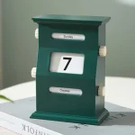 Relógio de calendário de mesa de madeira verde com dias e meses ajustáveis, colocado sobre um livro branco, com fundo neutro.