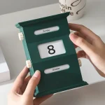 Calendário de mesa com dias, data e mês, em cores verde e branco, sendo manuseado por duas mãos.