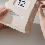 Calendário de mesa de madeira com data e mês ajustáveis, mostrando o dia 12 de novembro, com mãos ajustando a data.