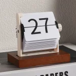 Calendário de mesa com números grandes em cartão rotativo, apoiado numa base de madeira, colocado numa superfície plana junto a uma parede texturizada.