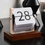Calendário de mesa vintage com números grandes e mecanismo de rotação, colocado numa base de madeira escura, com objetos decorativos ao fundo.