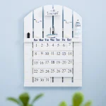 Calendário de parede com design náutico, incluindo barcos, faróis e elementos marítimos, com espaço para marcar dias e meses.