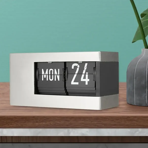 Relógio de mesa com calendário rotativo mostrando segunda-feira, 24 de outubro, ao lado de uma planta em vaso cinza escuro.