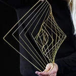 Escultura de fios dourados formando uma espiral geométrica, segurada por uma pessoa com roupa escura, fundo preto.
