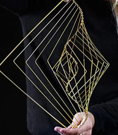 Escultura de fios dourados formando uma espiral geométrica, segurada por uma pessoa com roupa escura, fundo preto.