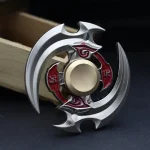 Fidget spinner com lâminas de metal prateado e detalhes vermelhos, com um centro dourado, colocado sobre uma superfície escura.