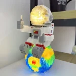 Estátua de astronauta feita com blocos de LEGO, com capacete iluminado e base colorida que simula uma explosão de cores.