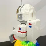 Estátua de Lego de um astronauta com capacete dourado, traje branco com detalhes em vermelho e azul, segurando uma ferramenta cinzenta, sobre uma base colorida.