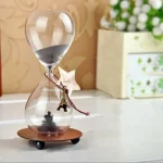 Relógio de areia decorativo com estrela de metal pendurada, base de madeira e areia cinzenta, colocado numa superfície branca com fundo de papel de parede floral.