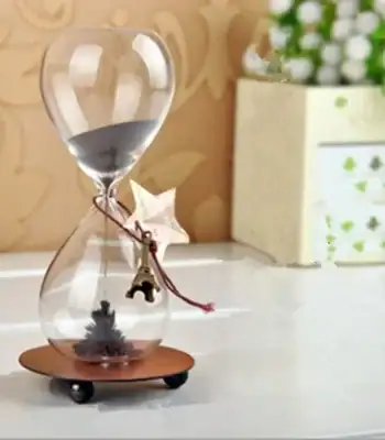 Relógio de areia decorativo com estrela de metal pendurada, base de madeira e areia cinzenta, colocado numa superfície branca com fundo de papel de parede floral.
