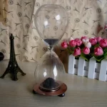 Ampulheta decorativa com areia preta, base de metal e vidro transparente, colocada numa mesa com decoração de flores e miniatura da Torre Eiffel ao fundo.