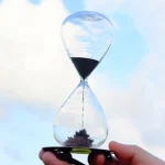 Relógio de areia transparente com areia preta a cair, mãos a segurar na base, céu azul ao fundo.